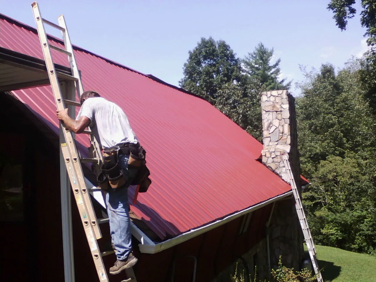 Roofer on a ladder beside a red metal roof and stone chimney — Soffit & Fascia Repair by Roofing Brothers in Elliott